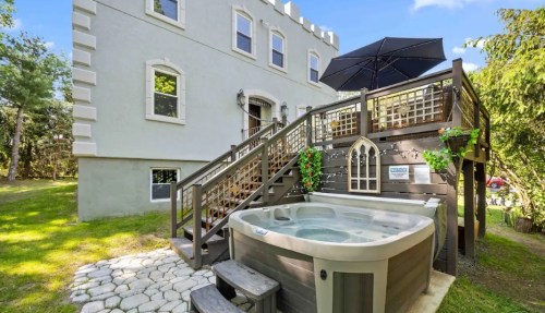 A hot tub on a stone patio next to a two-story house with a deck and an umbrella, surrounded by greenery.