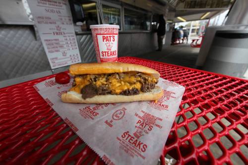 A cheesesteak sandwich with melted cheese on a hoagie roll, served with a drink on a red table.