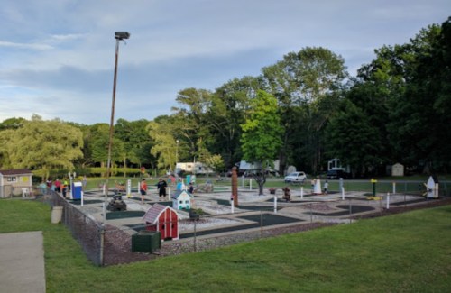 A miniature golf course with colorful obstacles, surrounded by trees and a grassy area, under a partly cloudy sky.