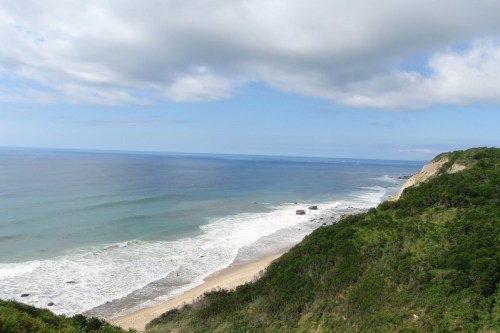 A scenic view of a coastline with waves crashing on the shore, surrounded by lush greenery and a cloudy sky.