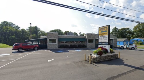 A restaurant with a stone exterior and large windows, featuring a sign and parked cars in the foreground.