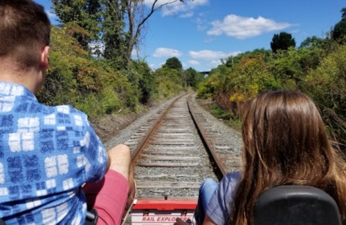 Two people sit on a rail cart, facing a long stretch of railway tracks surrounded by greenery under a blue sky.