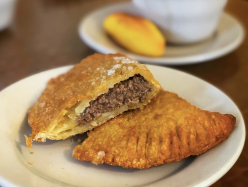 A golden-brown pastry filled with ground meat, served on a plate with a cup and a small pastry in the background.