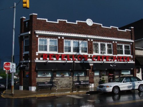 Exterior of Revello's Cafe Pizza, a brick building with large windows and a red sign, under a cloudy sky.