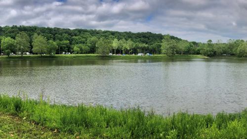A serene lake surrounded by lush greenery and trees, with a cloudy sky above and camping sites in the distance.