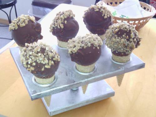 A display of six ice cream cones topped with chocolate and nuts, arranged on a metal stand.
