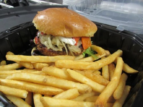 A burger with toppings on a bun served with a side of golden French fries in a black takeout container.