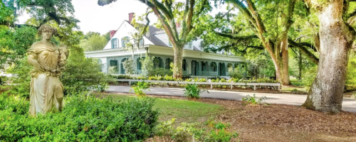 A historic house surrounded by lush greenery and a statue in the foreground, set in a tranquil garden.