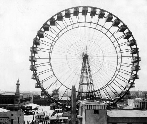 Historic black-and-white photo of a large Ferris wheel with surrounding buildings and a bustling fairground below.