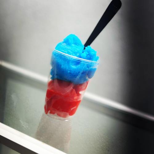 A colorful dessert in a clear cup, featuring layers of blue and red slushy ice with a black spoon.