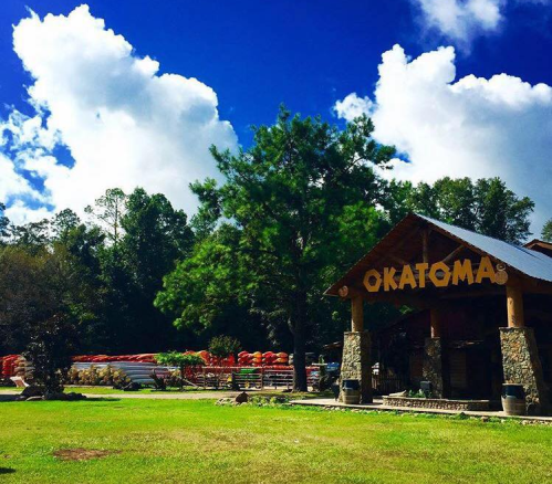 A rustic lodge with "OKATOMA" sign, surrounded by trees and blue skies, with kayaks visible in the background.