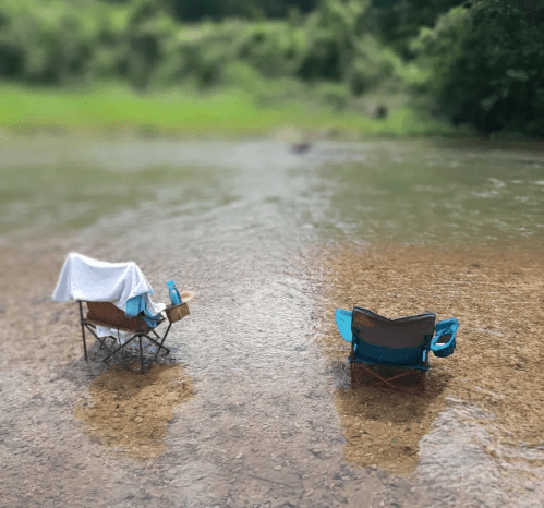 Two camping chairs sit in shallow water, surrounded by greenery, with a towel draped over one chair.