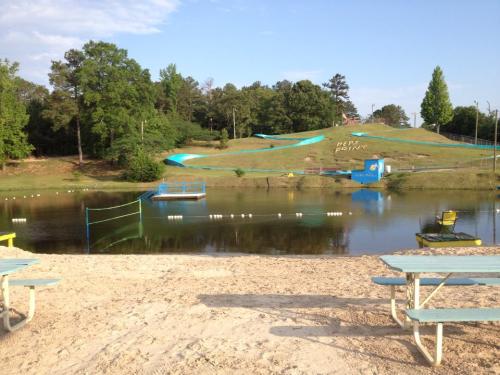 A sandy beach area by a calm lake, with green trees and a water slide in the background. Picnic tables are visible.