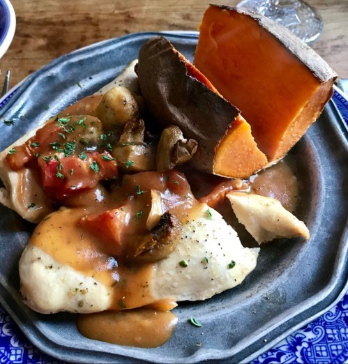 A plate of tender chicken with gravy, roasted vegetables, and a baked sweet potato, garnished with herbs.