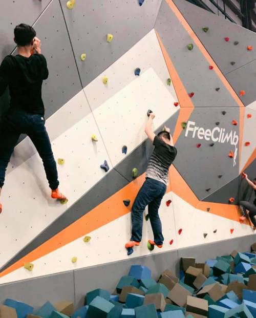 Two climbers ascend a colorful indoor rock wall, with blue climbing holds and a padded floor below.