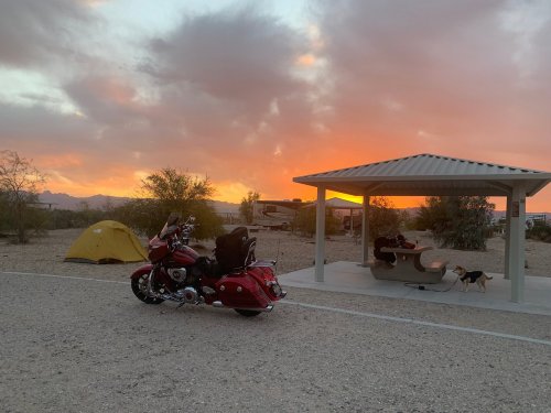 A motorcycle parked near a tent and picnic area, with a vibrant sunset and desert landscape in the background.