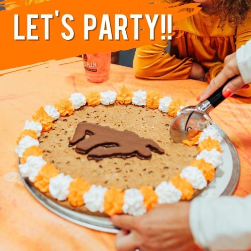 A decorated cookie cake with orange and white frosting, featuring a playful design, surrounded by festive party decorations.