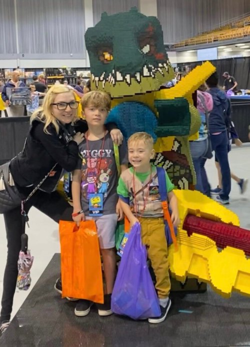 A woman and two children pose with a large LEGO dinosaur sculpture at an event, each holding colorful bags.