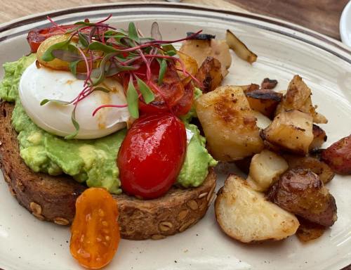 Avocado toast topped with a poached egg, cherry tomatoes, microgreens, and a side of roasted potatoes.