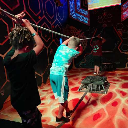 Two children pull on ropes in a colorful, interactive play area with a honeycomb design on the walls.
