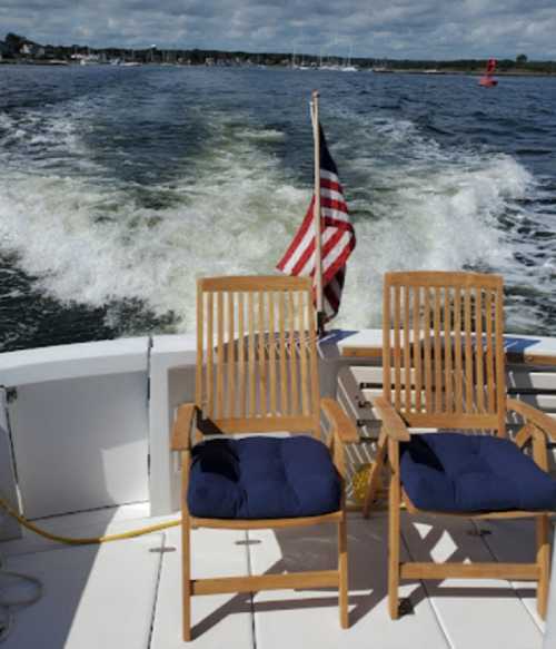 Two wooden chairs with blue cushions on a boat, with an American flag waving and a scenic water view behind.