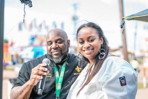 A man and woman smile together at an outdoor event, with the man holding a microphone and wearing a chef's jacket.