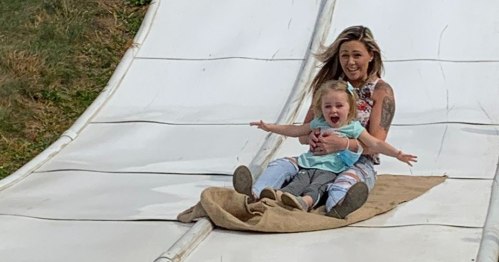 A woman and a young child joyfully slide down a smooth, white slide, both smiling and having fun.