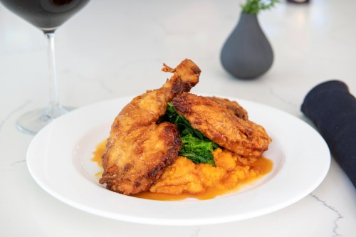 A plate of crispy fried chicken served on a bed of sweet potato mash, garnished with greens and a glass of red wine.