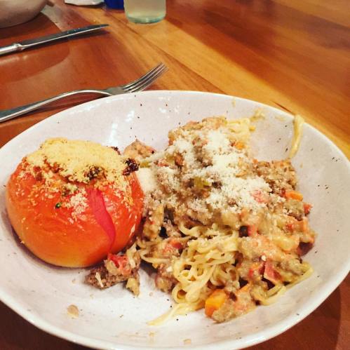 A bowl of pasta with meat sauce and a stuffed tomato, garnished with grated cheese, on a wooden table.