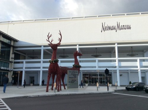 Two large red reindeer sculptures adorned with greenery stand outside a Neiman Marcus store entrance.