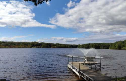 A serene lake view with a dock and fountain, surrounded by trees under a partly cloudy sky.