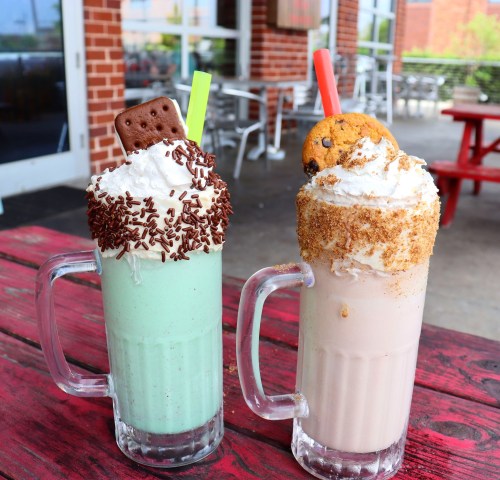Two colorful milkshakes topped with whipped cream, sprinkles, and cookies, served in clear mugs on a red table.