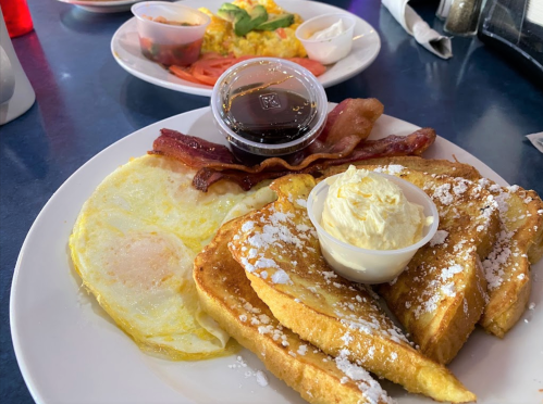 A plate of French toast with powdered sugar, bacon, a fried egg, and a small cup of syrup and butter.