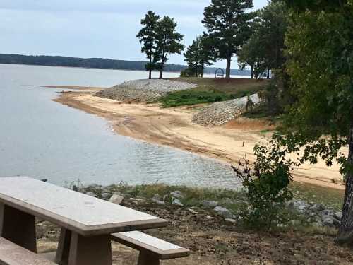 A sandy shoreline by a calm lake, with trees and a picnic table in the foreground under a cloudy sky.