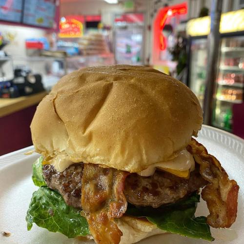 A close-up of a juicy burger with bacon, lettuce, and cheese on a bun, served on a plate in a diner setting.