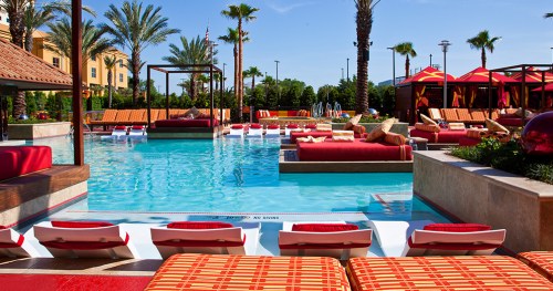 A vibrant pool area with lounge chairs, cabanas, and palm trees under a clear blue sky.
