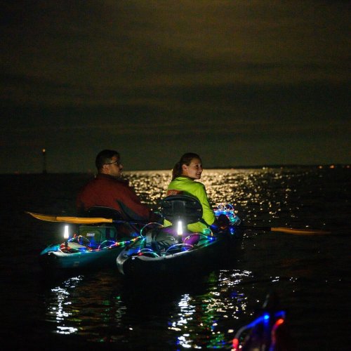 Two people in kayaks with glowing lights paddling on a dark water surface under a night sky.