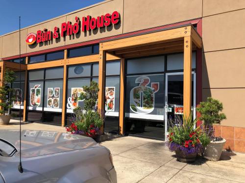 Exterior of a restaurant named "Bánh & Phở House," featuring large windows and colorful flower planters.