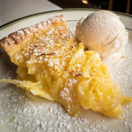 A slice of lemon pie dusted with powdered sugar, served with a scoop of vanilla ice cream on a white plate.