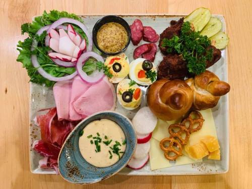 A colorful platter featuring meats, cheeses, pickles, radishes, pretzels, and deviled eggs, garnished with greens.