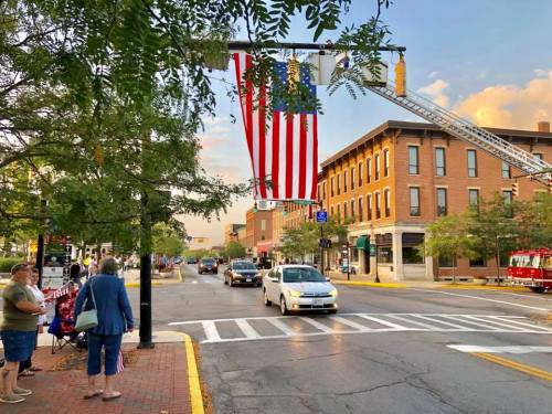 A street scene with a large American flag hanging from a fire truck ladder, people walking, and cars passing by.