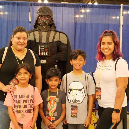 A group of five people, including two adults and three children, pose in front of a large LEGO Darth Vader statue.