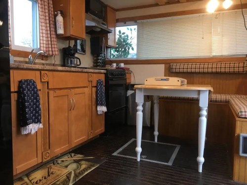 Cozy kitchen interior with wooden cabinets, a small table, and a seating area, featuring a stove and coffee maker.