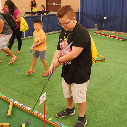 A child plays mini-golf indoors, focusing on their shot while another child walks in the background.