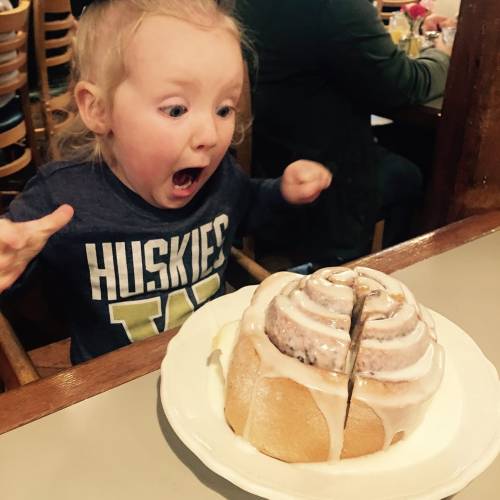 A surprised child with wide eyes excitedly looks at a large cinnamon roll on a plate.