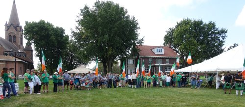 A crowd of people in green attire holding Irish flags at an outdoor event with tents and a church in the background.