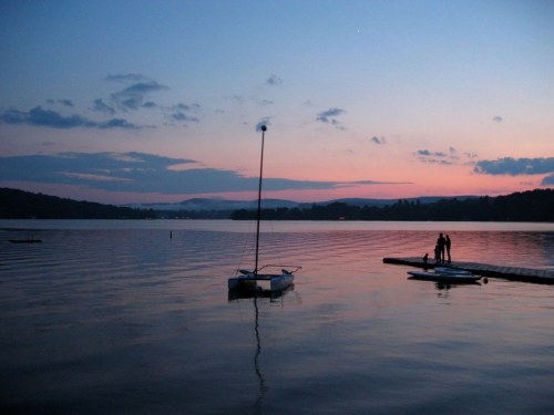 A serene lake at sunset, with a sailboat and two people standing on a dock, surrounded by calm waters and hills.