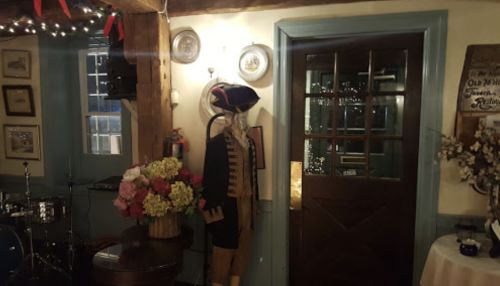 A historical figure mannequin in a colonial outfit stands beside a door in a cozy, decorated room.