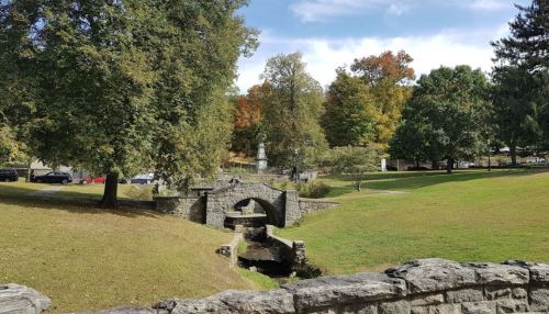 A stone bridge over a small stream in a park with trees displaying autumn colors and a statue in the background.