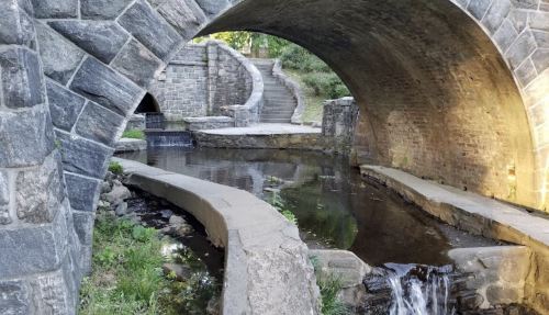A serene stone bridge arches over a calm stream, with steps leading up to a pathway in a lush, green setting.